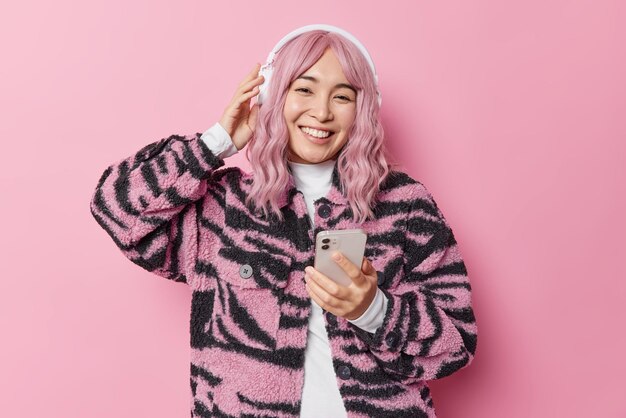 Una mujer asiática bastante joven con el pelo teñido escucha música a través de auriculares estéreo sostiene un teléfono móvil vestido con una chaqueta de invierno y tiene buen humor aislado sobre un fondo rosado. Concepto de personas y pasatiempos.