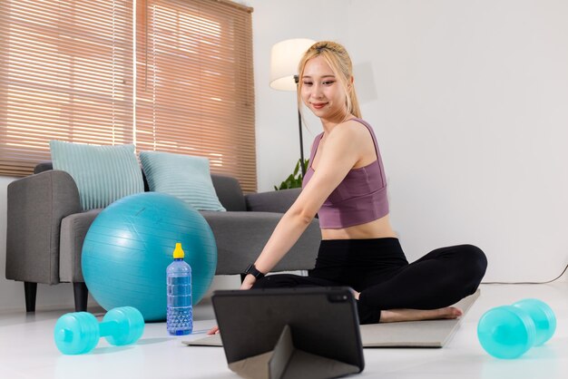 Mujer asiática bastante joven en ropa deportiva sentada en una alfombra de ejercicio y viendo videos de entrenamiento en línea en una tableta digital