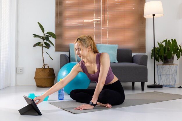 Mujer asiática bastante joven en ropa deportiva sentada en una alfombra de ejercicio y viendo videos de entrenamiento en línea en una tableta digital