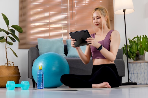 Mujer asiática bastante joven en ropa deportiva sentada en una alfombra de ejercicio y viendo videos de entrenamiento en línea en una tableta digital
