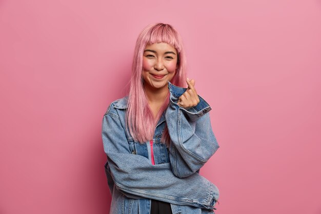 Mujer asiática bastante romántica con linda sonrisa de amor, cabello largo de color rosa, hace el signo del dedo del corazón coreano, expresa amor y simpatía, confesión