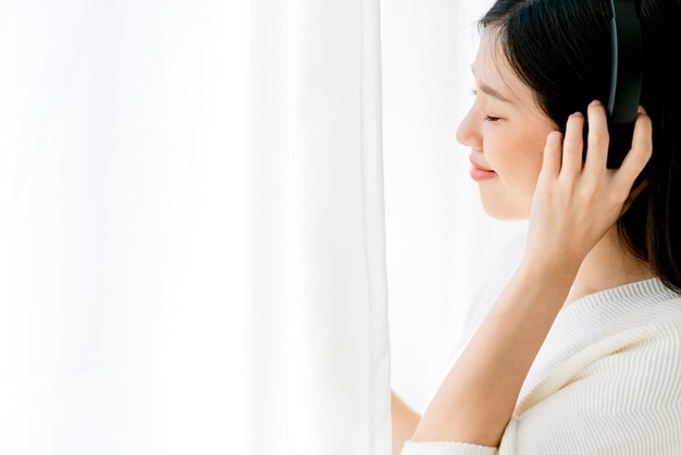 La mujer asiática de la belleza de la felicidad disfruta de la música de la canción con el medio de los auriculares en el fondo de la cortina blanca de la sala de estar