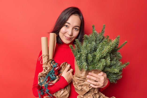 Una mujer asiática bonita y complacida con el pelo oscuro lleva atributos navideños para la decoración espera el Año Nuevo vestida con un cuello de tortuga casual aislado sobre una pared roja. Encantadora modelo femenina se prepara para Navidad