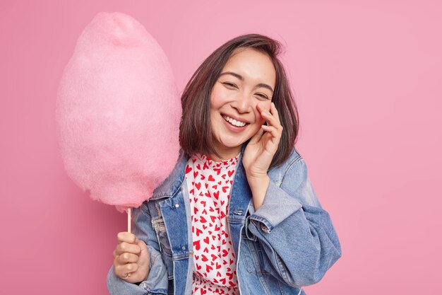 Mujer asiática bonita despreocupada positiva sonríe sinceramente disfruta del tiempo libre come dulce algodón de azúcar vestido con chaqueta de mezclilla