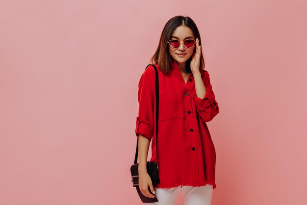 Una mujer asiática bronceada con una camisa roja de gran tamaño se pone gafas de sol y posa con un bolso en un fondo rosa