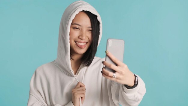 Mujer asiática con capucha sacando la lengua mientras toma un lindo selfie en el teléfono inteligente sobre fondo azul