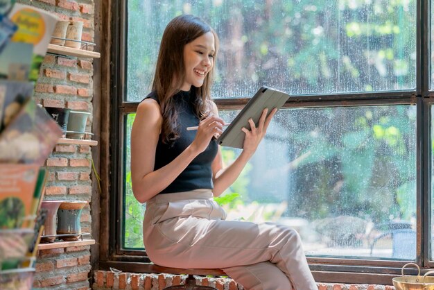 Una mujer asiática casual se relaja con la mano usando una tableta, navega por Internet, escribe un diario, sonríe alegremente mientras se sienta en el cálido interior de un café cerca de una gran ventana.