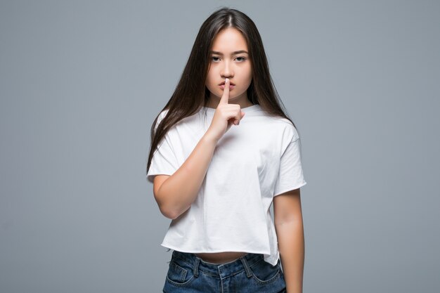 Mujer asiática casual sonriente que muestra gesto del silencio con el dedo sobre sus labios sobre fondo gris