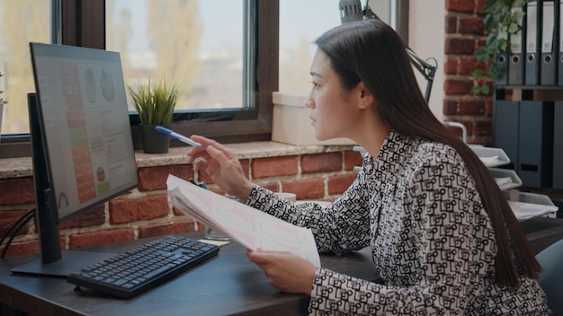 Mujer asiática comparando gráficos en papeles y computadoras, trabajando para planificar un proyecto de negocios en la oficina de inicio. Empleado de la empresa analizando documentos para diseñar datos financieros y estrategia.