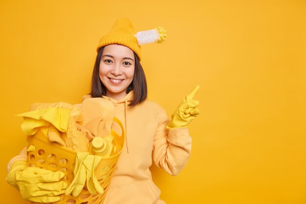 La mujer asiática complacida lava la ropa en casa usa un sombrero con una sudadera con capucha de cepillo de baño atascado y guantes de goma apunta a un espacio de copia en blanco aislado sobre fondo amarillo que muestra el producto para limpiar
