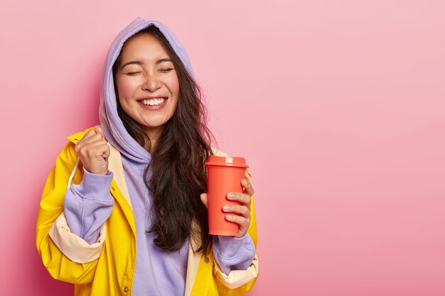 La mujer asiática complacida y llena de alegría levanta el puño cerrado, mantiene los ojos cerrados, bebe la bebida caliente del frasco, se regocija en el momento feliz, usa ropa abrigada impermeable