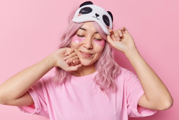 Una mujer asiática complacida con el pelo rosado aplica una máscara cosmética para los ojos por tener una piel sana y fresca que mantiene los ojos cerrados, usa una venda en los ojos y una camiseta informal aislada sobre un fondo rosa Concepto de belleza