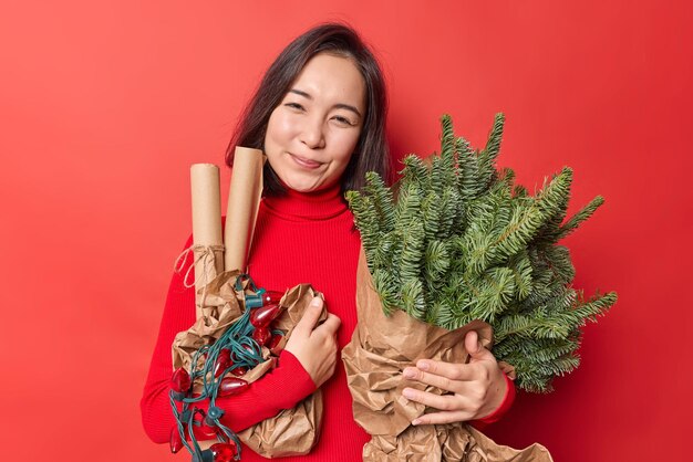 La mujer asiática complacida sonríe alegremente sostiene artículos de vacaciones para la decoración anticipada para el Año Nuevo y la Navidad usa poses de cuello alto contra un fondo rojo vivo. Concepto de celebración y preparación.