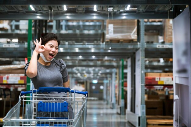 Mujer asiática de compras de seguridad con máscara facial gesto de mano de protección hacer una señal de estar bien para expresar una emoción segura mientras tira del carrito de compras revisando elegir artículos en las compras del estante del almacén