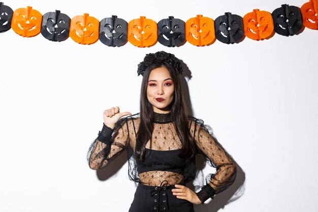 Mujer asiática confiada en elegante vestido gótico y corona negra apuntando a sí misma como profesional, de pie contra las decoraciones de halloween.