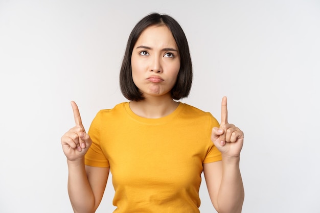 Mujer asiática decepcionada que mira señalar con el dedo hacia arriba con expresión de cara malhumorada enojada de pie en una camiseta amarilla sobre fondo blanco