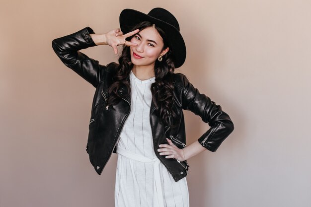 Mujer asiática despreocupada posando con el signo de la paz. Foto de estudio de feliz mujer coreana lleva elegante sombrero y chaqueta de cuero.