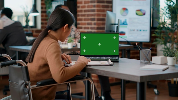 Mujer asiática con discapacidad usando pantalla verde en una laptop, trabajando con cromakey aislado y plantilla de maqueta. Persona en silla de ruedas mirando el fondo del espacio de copias en blanco en la pantalla.