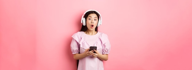 Una mujer asiática emocionada con auriculares inalámbricos dice wow sosteniendo un teléfono móvil y mirando divertida a la cámara