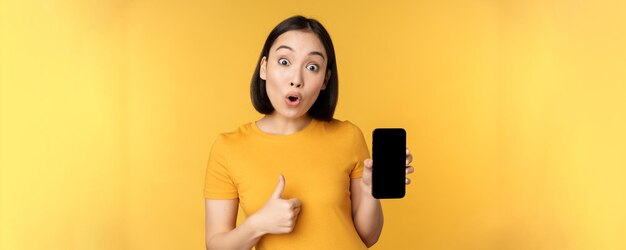 Mujer asiática emocionada que muestra la pantalla del teléfono móvil con los pulgares hacia arriba como algo bueno recomendando la aplicación del teléfono inteligente de pie sobre un fondo amarillo