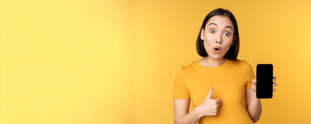 Mujer asiática emocionada que muestra la pantalla del teléfono móvil con los pulgares hacia arriba como algo bueno recomendando la aplicación del teléfono inteligente...