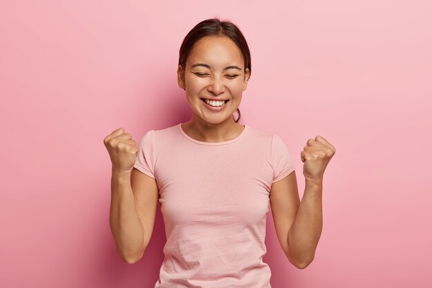 La mujer asiática emocional exitosa aprieta los puños en el aire, celebra el éxito, tiene una amplia sonrisa, mantiene los ojos cerrados, está emocionada y alegre, disfruta del dulce sabor de la victoria, usa ropa informal.