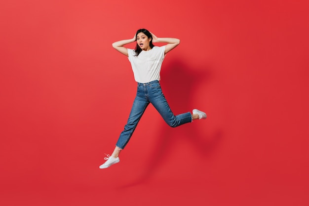 Mujer asiática emocional en jeans saltando sobre pared roja