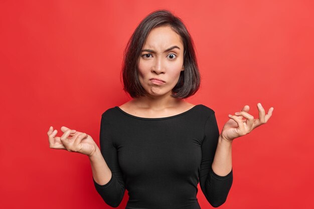 La mujer asiática enojada levanta las cejas de la molestia mantiene las palmas de las manos hacia los lados se encoge de hombros con vacilación viste un jersey negro aislado sobre una pared roja vívida que se ve con desconcierto.