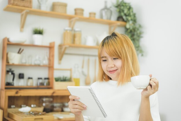 La mujer asiática escribe listas de compras en la libreta por la pluma en su contador de la cocina en casa y leyendo