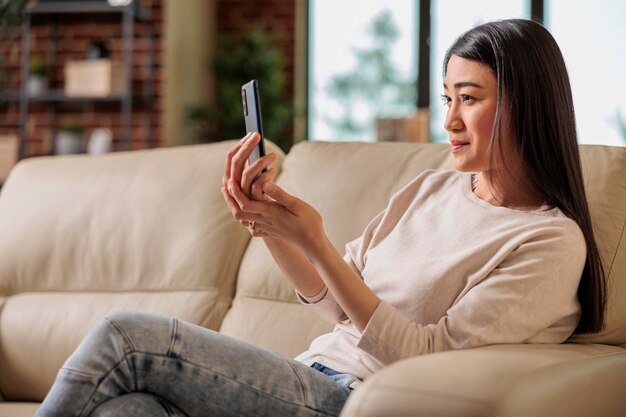 Mujer asiática feliz de aspecto auténtico casual que usa un teléfono inteligente en casa, tecnología de Internet web usuario de teléfono inteligente mensaje móvil dispositivo digital de comunicación de redes sociales