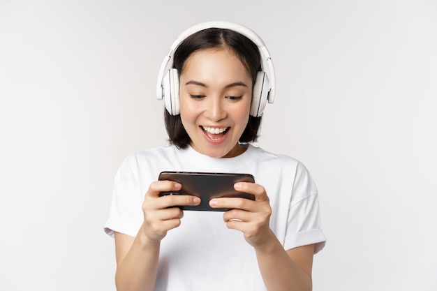 Mujer asiática feliz en auriculares mirando el teléfono inteligente viendo videos en el teléfono móvil y sonriendo de pie sobre fondo blanco