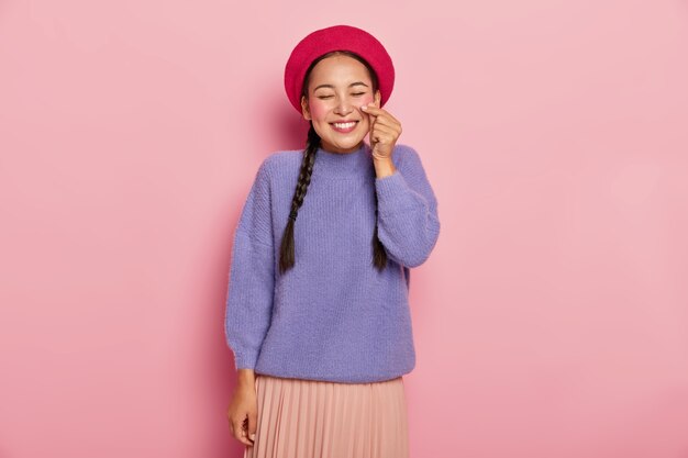 La mujer asiática feliz y complacida da forma a un pequeño corazón con las manos, hace que el coreano sea como un signo, usa boina roja, suéter casual y falda, sonríe agradablemente, está de buen humor, aislado sobre una pared rosa