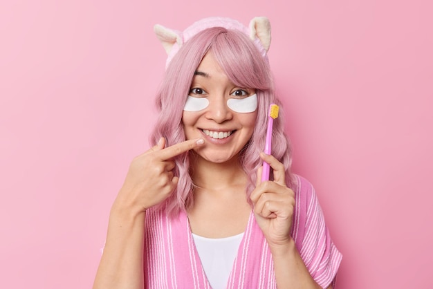 Una mujer asiática feliz con puntos de cabello rosado en una sonrisa con dientes sostiene un cepillo de dientes que se somete a procedimientos de belleza e higiene y usa ropa informal posa en el interior contra un fondo rosado. Mira mis dientes blancos