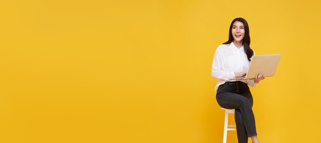 mujer asiática feliz sonriendo mientras ella usa una computadora portátil sentada en una silla blanca y mirando el espacio de la copia