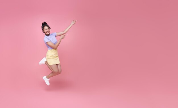 Mujer asiática feliz sonriendo y saltando mientras las manos apuntan aisladas sobre el espacio de copia de fondo rosa