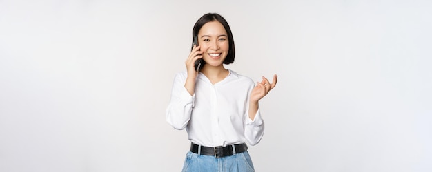Mujer asiática feliz sonriente hablando por teléfono inteligente con la vendedora del cliente de guardia sosteniendo el teléfono móvil y gesticulando de pie sobre fondo blanco
