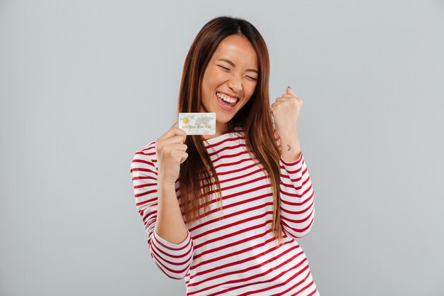 La mujer asiática feliz en suéter se regocija y que sostiene la tarjeta de crédito sobre fondo gris