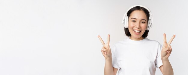 Mujer asiática feliz usando auriculares y sonriendo mostrando paz vssign escuchando música de pie en tsh