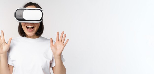 Mujer asiática feliz usando auriculares VR agitando las manos levantadas y riendo usando gafas de realidad virtual de pie sobre fondo blanco.