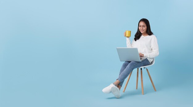 mujer asiática feliz usando computadora portátil sentada en una silla blanca y tomando café