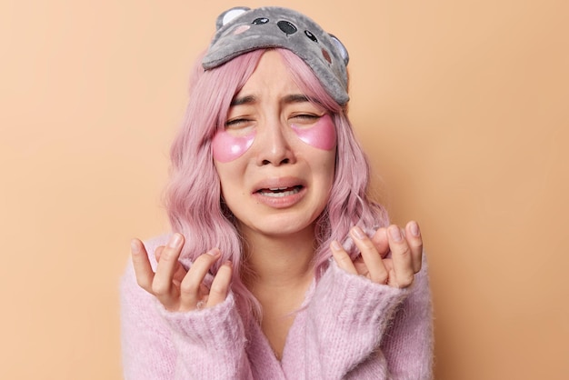 Una mujer asiática frustrada y abatida llora de desesperación y aplica parches de hidrogel debajo de los ojos, tiene el pelo largo y rosado, usa un puente y una máscara para dormir en la frente aislada sobre un fondo marrón Concepto de belleza