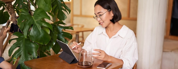 Foto gratuita mujer asiática con gafas viendo algo en una tableta digital tomando café en una cafetería y sonriendo trabajando