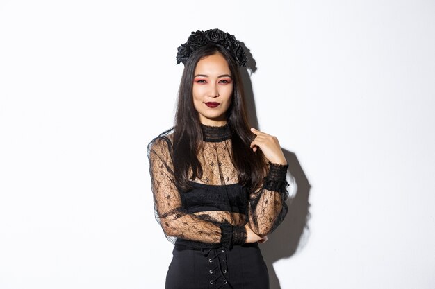 Mujer asiática hermosa y atrevida vestida con vestido de encaje negro y corona para la fiesta de halloween. Mujer con maquillaje gótico sonriendo complacida, mirando a la cámara confiada.