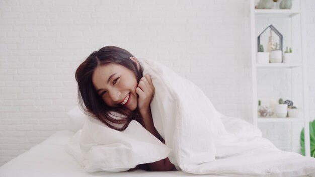 La mujer asiática hermosa feliz despierta, sonriendo y estirando sus brazos en su cama en el dormitorio.