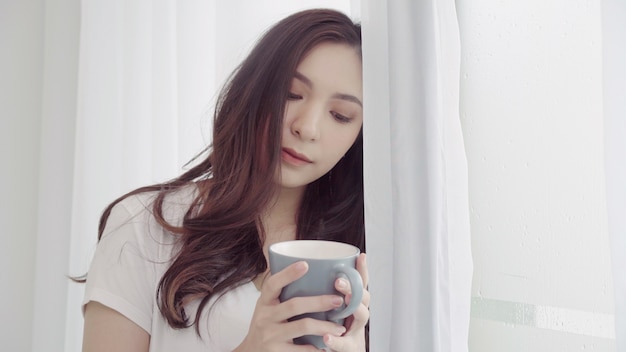 Mujer asiática hermosa feliz que sonríe y que bebe una taza de café o de té cerca de la ventana en el dormitorio.