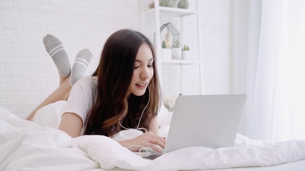 Mujer asiática hermosa que juega el ordenador mientras que miente en la cama en su dormitorio.