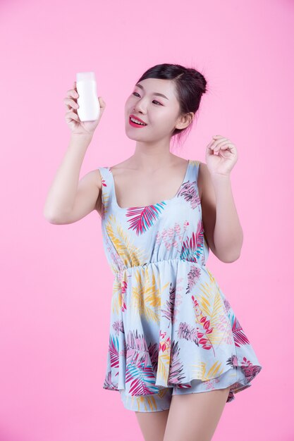 Mujer asiática hermosa que sostiene una botella de producto en un fondo rosado.