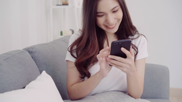 Mujer asiática hermosa que usa smartphone mientras que miente en el sofá en su sala de estar.