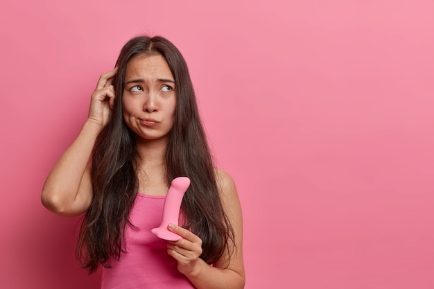 Una mujer asiática indecisa se frota la cabeza elige un consolador en un sex shop sostiene un objeto fálico rosado de silicona para proporcionar placer sexual a través de la penetración encuentra el tamaño perfecto para ella, subsititues genitales reales de los hombres