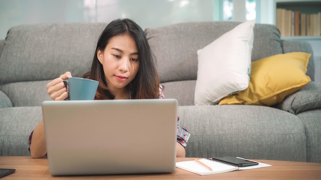 Mujer asiática independiente joven del negocio que trabaja en la computadora portátil que comprueba medios sociales y que bebe el café mientras que miente en el sofá cuando se relaja en sala de estar en casa.